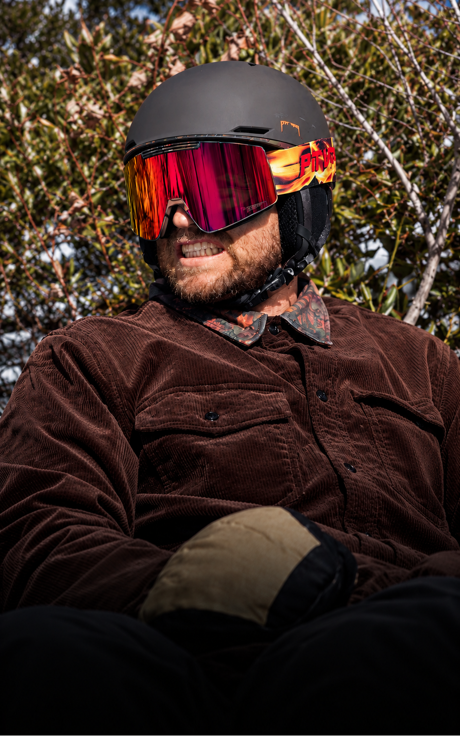 A man wearing a matte black ski helmet with vibrant red, orange, and yellow mirrored goggles, a brown corduroy jacket, and a tan and black winter glove, set against a background of outdoor trees.