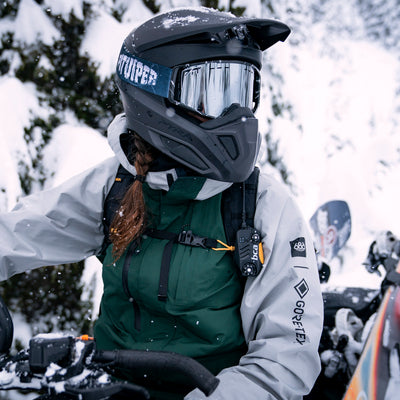 A person wearing a black full-face snowmobile helmet with a reflective visor, a dark green insulated jacket, and a light gray jacket, riding a snowmobile in a snowy outdoor setting.