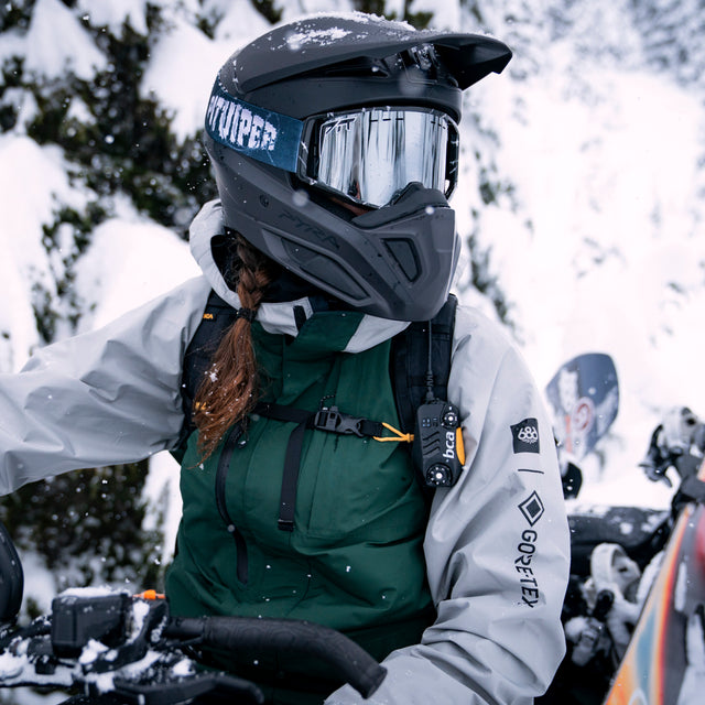 A person wearing a black full-face snowmobile helmet with a reflective visor, a dark green insulated jacket, and a light gray jacket, riding a snowmobile in a snowy outdoor setting.