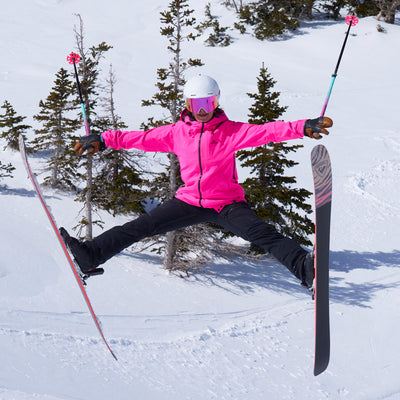 A skier jumps mid-air in a snowy mountain setting, wearing a bright pink ski jacket, black ski pants, a white helmet, and pink-tinted goggles, with ski poles and skis extended.