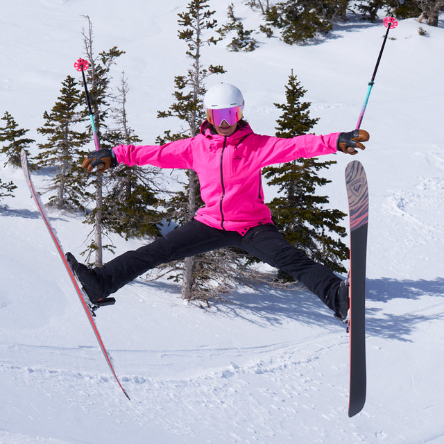 A skier jumps mid-air in a snowy mountain setting, wearing a bright pink ski jacket, black ski pants, a white helmet, and pink-tinted goggles, with ski poles and skis extended.