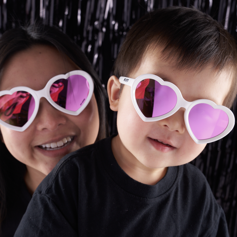 Pink | you_can_match_mom.JPG | A close-up lifestyle shot of a woman and a young child wearing white heart-shaped sunglasses with pink-tinted lenses against a black tinsel backdrop.