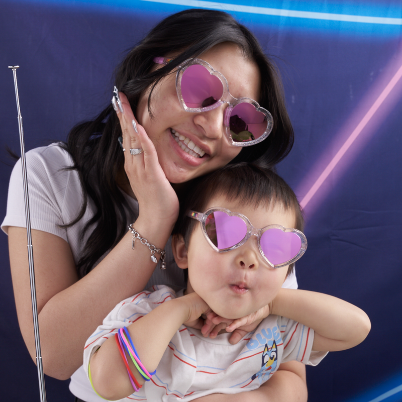 Pink | you_can_match.JPG | A smiling woman and a young child wearing heart-shaped pink sunglasses pose together in front of a blue photo booth backdrop with neon light effects. 