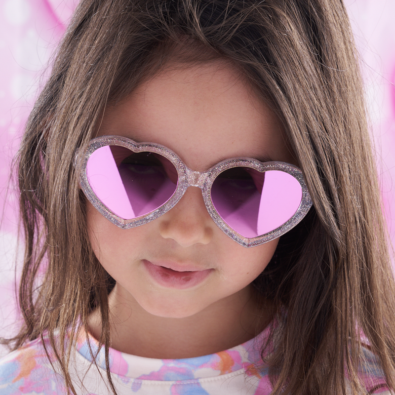 Pink | photo_of_person.JPG | A close-up of a young girl with long brown hair wearing heart-shaped sunglasses with glittery silver frames and pink mirrored lenses, set against a soft pink background