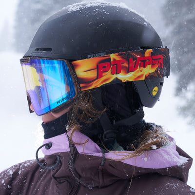 A person in a snowy outdoor setting wearing a black ski helmet, blue mirrored goggles with a red and orange flame-pattern strap, and a brown jacket.