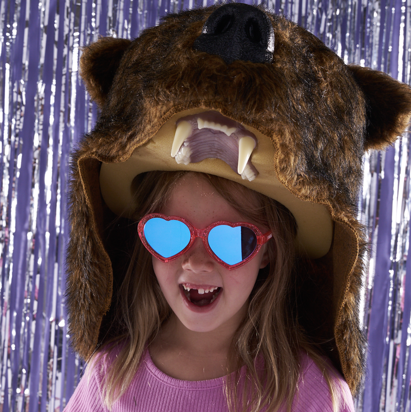 Blue-Purple | bear.JPG | A young girl wearing a brown bear costume hat with floppy ears, faux fur, a black snout, and large white teeth, paired with red glitter heart-shaped sunglasses with blue mirrored lenses, posed in front of a purple tinsel backdrop.