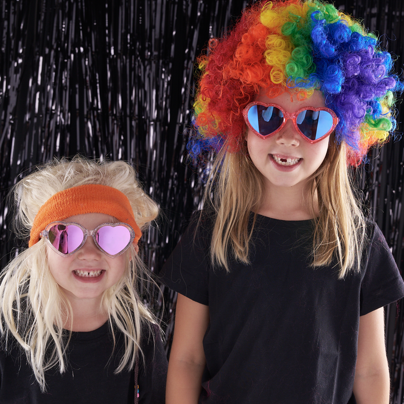 Blue-Purple | smile2x.JPG | Two children wearing colorful party accessories, including a rainbow clown wig, heart-shaped mirrored sunglasses, and an orange headband, posed against a black tinsel backdrop.