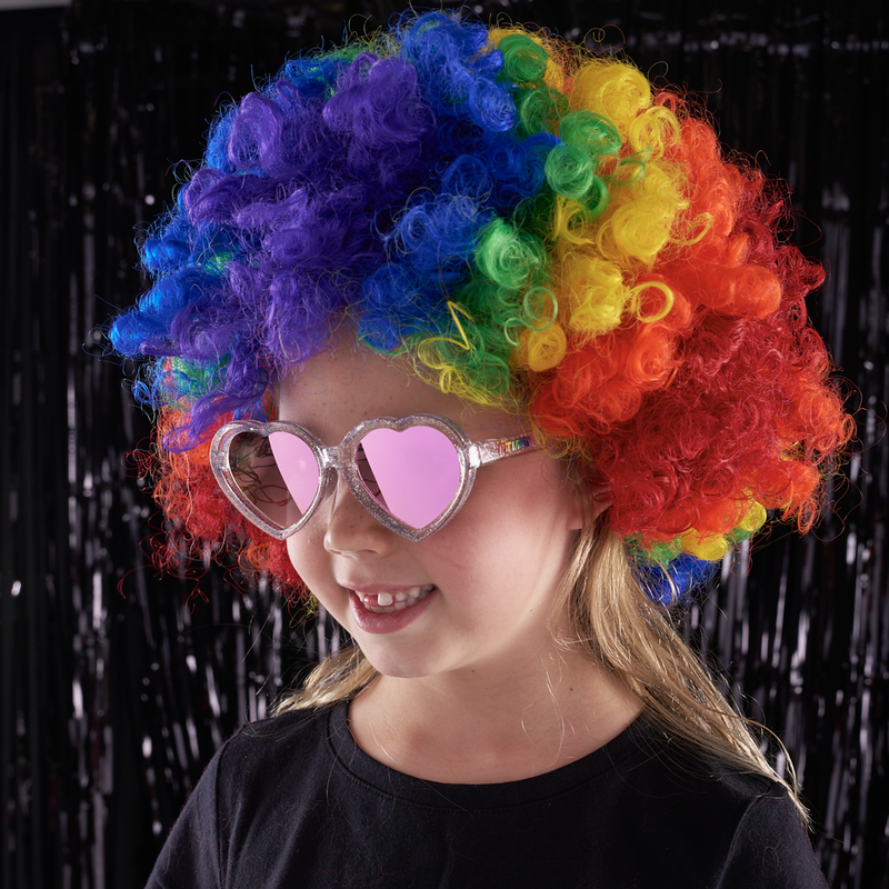 Pink | rainbow.JPG | A young girl wearing a vibrant rainbow-colored curly wig and heart-shaped pink sunglasses, posing against a black tinsel curtain background.
