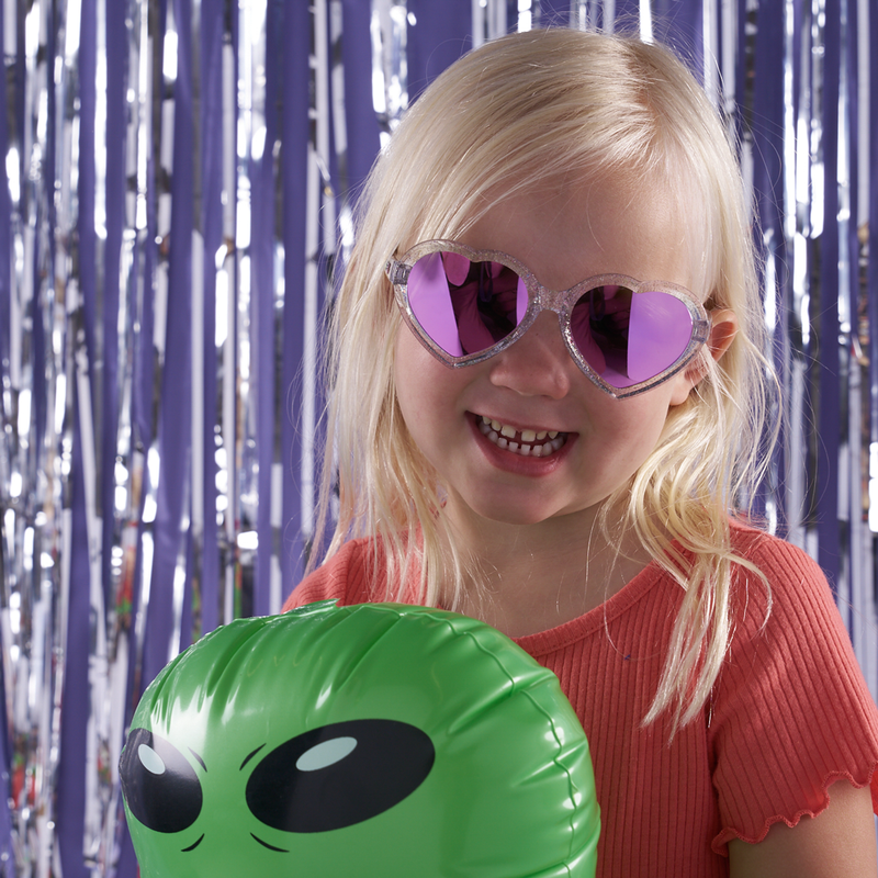 Pink | smile!.JPG | A young blonde girl wearing heart-shaped pink sunglasses and a coral pink top, smiling while holding a green inflatable alien head against a silver tinsel backdrop.