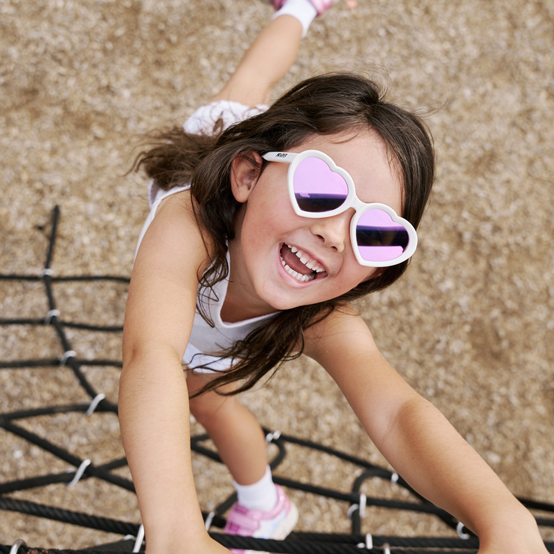 Pink | all_smiles.JPG | A young girl smiling and crawling on a playground rope net, wearing white heart-shaped sunglasses with purple mirrored lenses