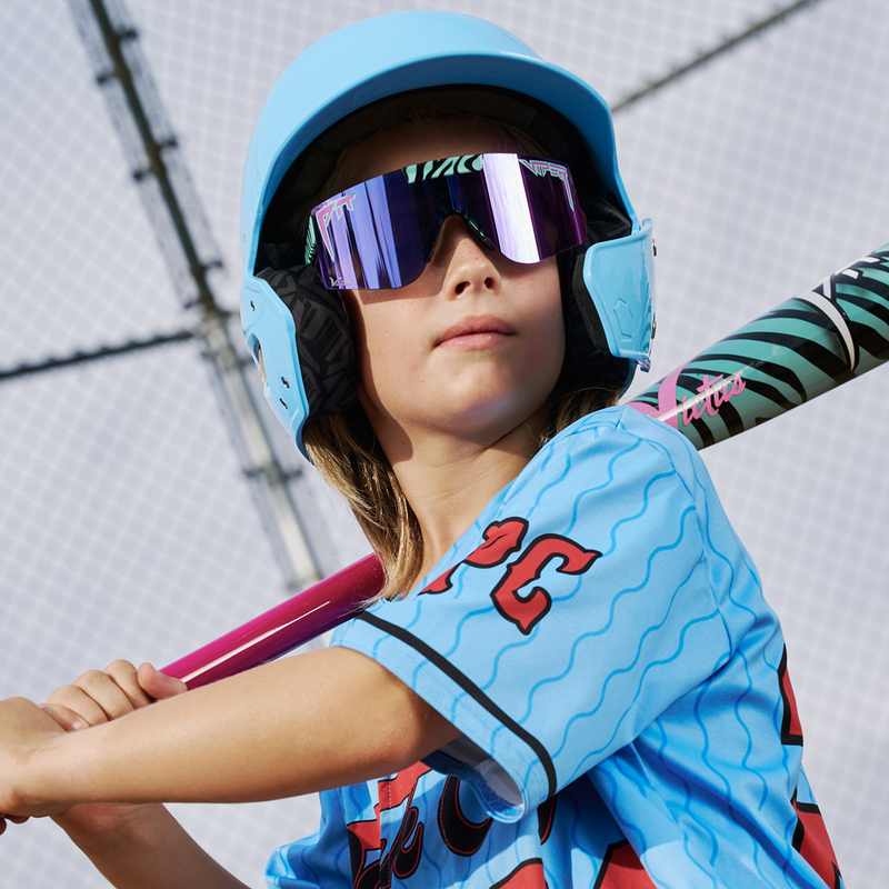 | batter_up.JPG | A young baseball player wearing a light blue helmet, mirrored purple sunglasses, and a light blue jersey with red accents, holding a pink bat. The background shows a chain-link fence.