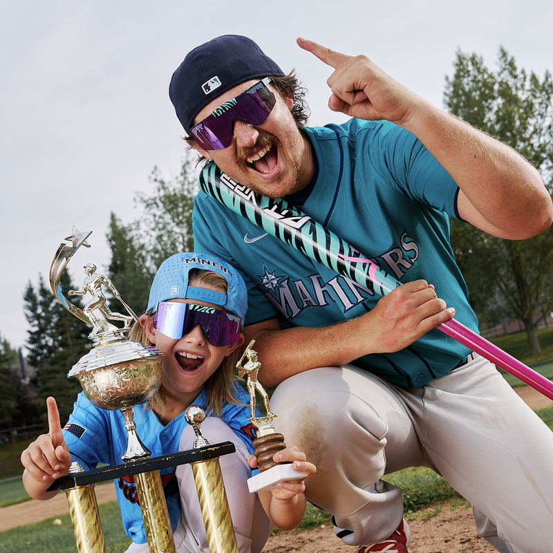 | we_are_number_1.JPG | A man and a child in baseball uniforms celebrate with trophies on a field. The man wears a teal  jersey, white pants, a navy cap, and purple-tinted sunglasses, while the child wears a matching blue cap and light blue jersey.