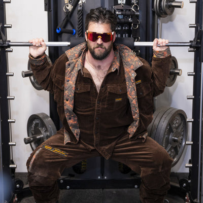 A man in a brown corduroy tracksuit and sunglasses squats inside a weight machine gym setting.