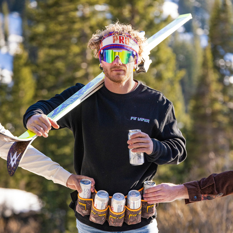 | man_holding_only_skis.JPG | A man outdoors in a snowy mountain setting, wearing a black sweatshirt with 'FIT VAPOR' on the chest, blue denim shorts, and holding a ski over his shoulder. He is surrounded by hands holding energy drinks and a can, with pine trees in the background.