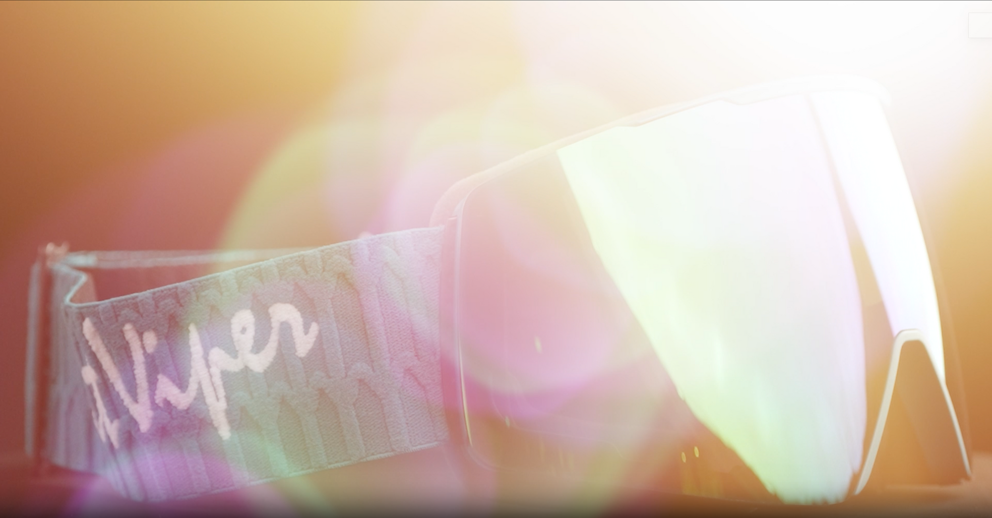 A pair of shield-style ski goggles with a clear lens and a patterned strap featuring the script brand name 'Viper' in white, shown against a warm, heavily filtered background with pink and yellow bokeh.
