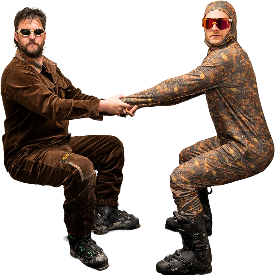 Two men in full-body camouflage ski suits and helmets pose against a solid white background, holding hands in a balancing pose. One man wears a solid brown velvet jumpsuit and sunglasses, while the other wears a brown and black camouflage ski suit with orange-tinted sunglasses.